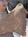 A person wearing glasses holds up a chunky knit triangular shawl in a warm beige/tan color with multicolored flecks and bright pink pom-pom trim along the edges.