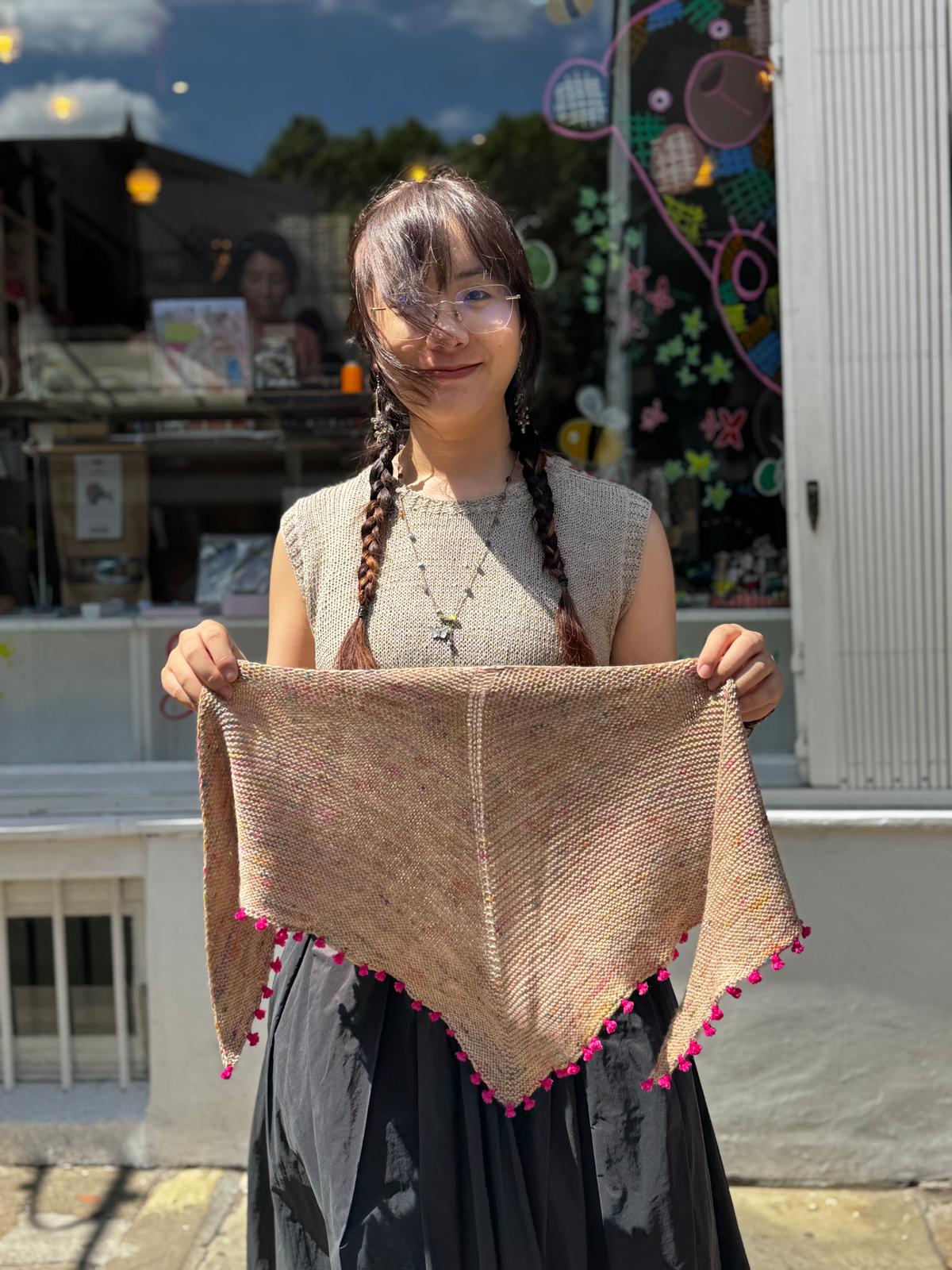 A woman with braided hair holds up a triangular knit shawl in a warm tan color with pink pom-pom trim, standing outdoors in front of a shop window.