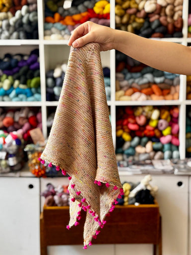 A hand holds up a small triangular beige knit scarf with multicolored yarn flecks and bright pink pom-pom trim, set against a blurred background of shelves filled with colorful yarn.