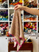 A hand holds up a small triangular beige knit scarf with multicolored yarn flecks and bright pink pom-pom trim, set against a blurred background of shelves filled with colorful yarn.