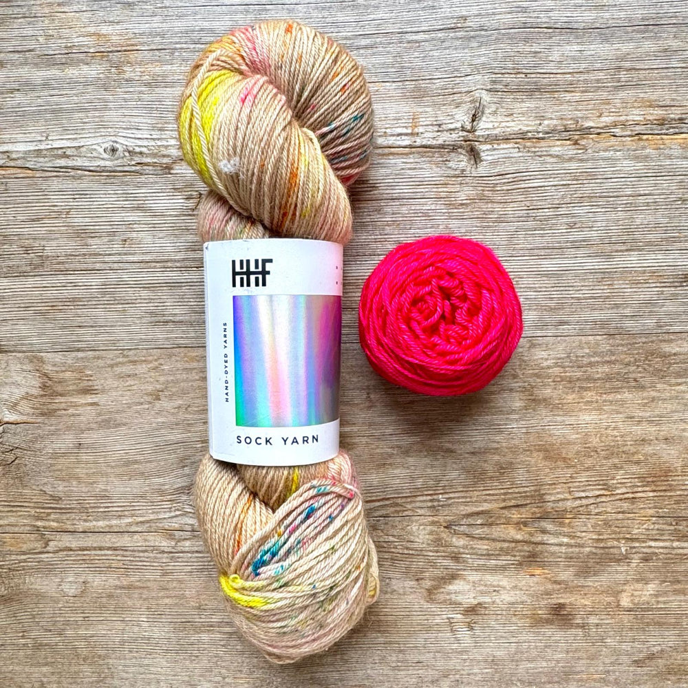 A skein of variegated sock yarn with a white paper label reading 'HUF SOCK YARN' sits on a rustic wooden surface next to a bright pink ball of yarn.