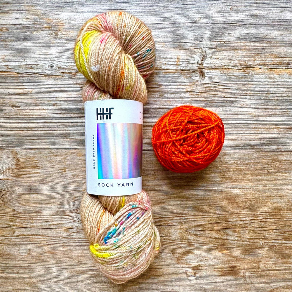Two skeins of yarn on a rustic wooden surface: one variegated pastel skein with yellow, pink, and blue flecks, and one bright orange-red skein. The variegated skein has a white paper label reading 'HHF SOCK YARN'.
