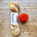 Two skeins of yarn on a rustic wooden surface: one variegated pastel skein with yellow, pink, and blue flecks, and one bright orange-red skein. The variegated skein has a white paper label reading 'HHF SOCK YARN'.
