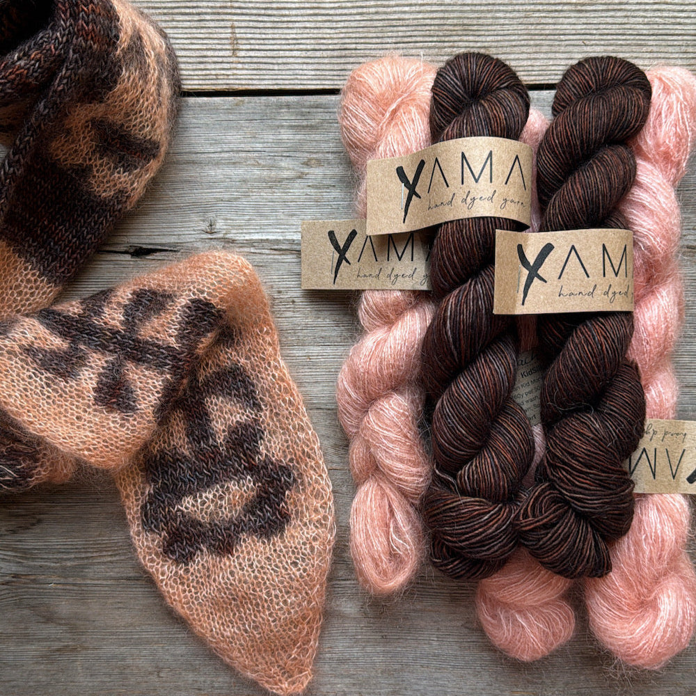 Three skeins of yarn in pink, brown, and black with 'YAMA' labels on a wooden surface.